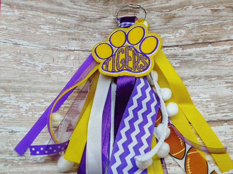 May include: A yellow and purple paw print keychain with the word "TIGERS" embroidered on it. The keychain has multiple ribbons in yellow, white, and purple, with white pom-poms attached.