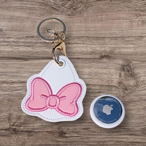 May include: A white faux leather keychain featuring an embroidered pink bow design with a gold-toned metal split ring and lobster clasp, accompanied by a circular white and blue electronic tracking device with a silver Apple logo on the front.