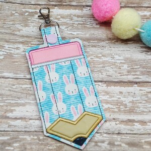 May include: A blue pencil-shaped keychain with a pink fabric rectangle and white bunny rabbit pattern. The keychain has a gold-colored pencil tip and a silver key ring.
