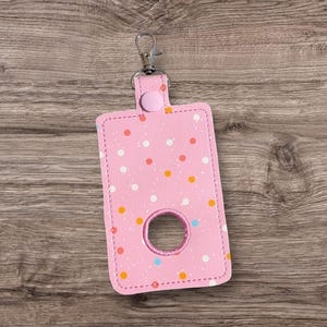 Puede incluir: Soporte rectangular rosa para desinfectante de manos con un clip plateado. El soporte presenta un estampado de lunares en blanco, naranja y azul. Se ve una abertura circular en el centro. El soporte está hecho de cuero de imitación cosido.