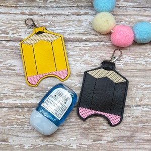May include: Two pencil-shaped hand sanitizer holders, one yellow and one black, with pink accents. Both holders have a key ring attached. A small blue hand sanitizer bottle is in front of the yellow holder.