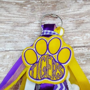 May include: A yellow paw print with the word "TIGERS" inside, attached to a purple, yellow, and white ribbon keychain.