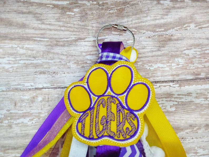 May include: A yellow paw print with the word "TIGERS" inside, attached to a purple, yellow, and white ribbon keychain.