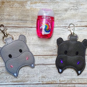 May include: Two gray and black hippopotamus-shaped hand sanitizer holders with keychains. The holders are made of faux leather and have embroidered eyes and mouths. The holders are attached to silver keychains.