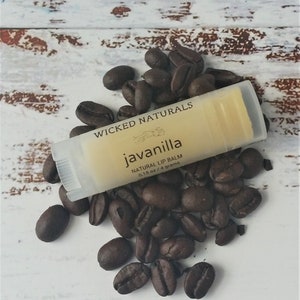 May include: A close-up of a small, clear tube of lip balm with a white label that reads "Wicked Naturals Javanilla Natural Lip Balm 0.15 oz / 4 grams". The tube is surrounded by coffee beans on a white wooden surface.