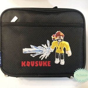 May include: Black rectangular Wildkin lunch bag with a zipper closure. The bag features a cartoon character with a yellow shirt and a blue ice-like weapon. The name "KOUSUKE" is printed in red.
