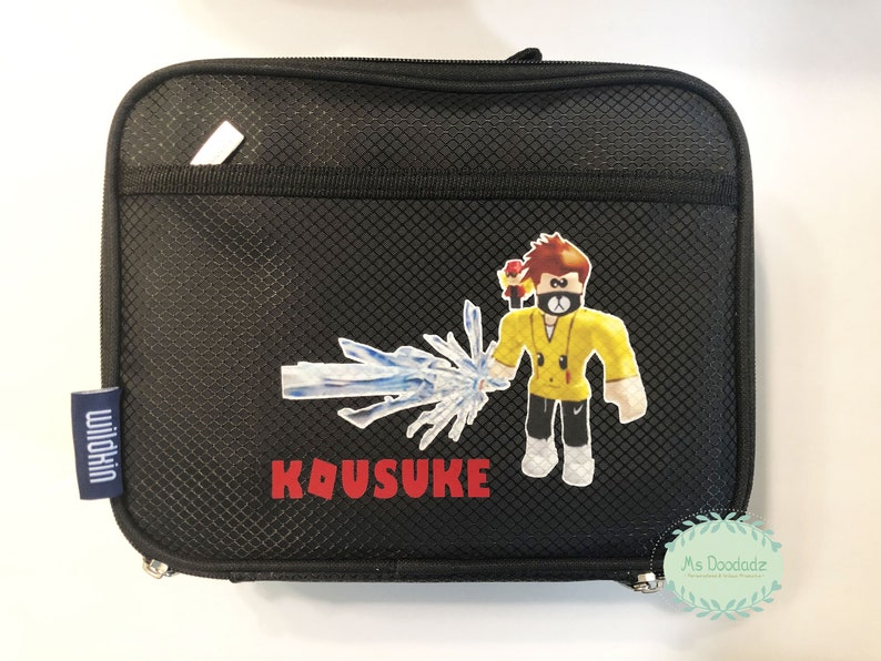 May include: Black rectangular Wildkin lunch bag with a zipper closure. The bag features a cartoon character with a yellow shirt and a blue ice-like weapon. The name "KOUSUKE" is printed in red.