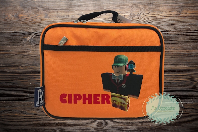 May include: Orange lunch bag with black trim and handle. Features a cartoon character with a green hat and the name "CIPHER" in red. A small item is in the front pocket. The bag has a zipper closure.