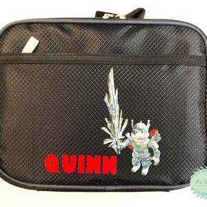 May include: Black zippered lunch bag with a textured pattern. A cartoon character with a sword and the name "QUINN" in red letters are on the front. The bag has a small pocket and a "Wildkin" tag.