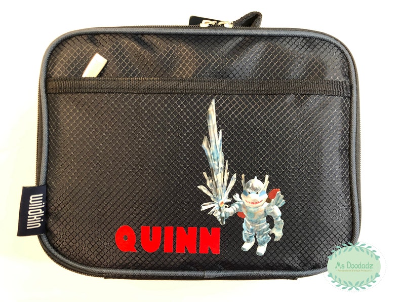 May include: Black zippered lunch bag with a textured pattern. A cartoon character with a sword and the name "QUINN" in red letters are on the front. The bag has a small pocket and a "Wildkin" tag.