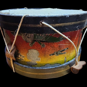 May include: A vintage toy drum with a colourful design featuring aeroplanes and clouds. The drum is made of wood and has a leather strap.