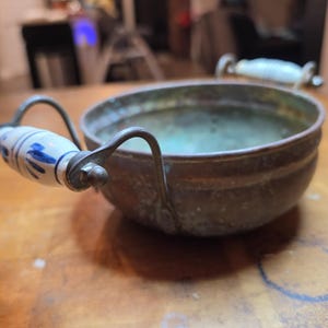 May include: A round, weathered copper bowl featuring two curved metal handles. Each handle is accented with a white ceramic grip decorated with blue floral patterns. The copper surface shows signs of oxidation and a dark, aged patina throughout.