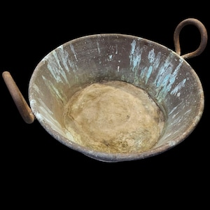 May include: An antique copper bowl with two handles. The bowl has a rounded shape with a wide opening and a flat bottom. The copper has a weathered appearance with areas of green oxidation, giving it a rustic look.