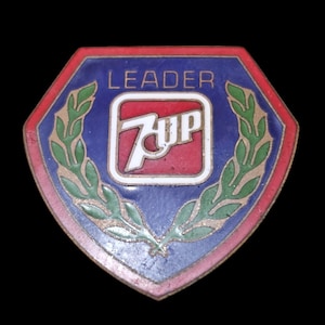 May include: A red, white, and blue enamel pin with the word "LEADER" above a red square with the word "7Up" in white. The pin is surrounded by a green laurel wreath.