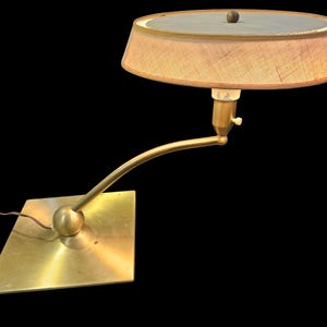 May include: A brass desk lamp with a curved arm and a round, off-white lampshade. The lamp has a square base.