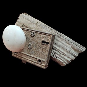 Könnte beinhalten: Ein antiker Türknauf mit einem weißen, runden Griff, einer rostigen Metallplatte und einem Stück verwittertem Holz. Die Metallplatte hat ein Schlüsselloch und Schraubendetails. Das Holz ist grau und strukturiert.