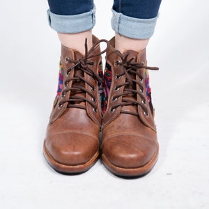May include: Brown leather ankle boots with colorful embroidered textile panels. The boots have a lace-up design and a rounded toe. The boots are worn with blue jeans.