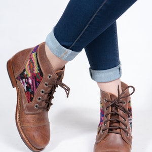 May include: Brown leather ankle boots with colorful woven textile panels. The boots have a lace-up design and are paired with dark blue denim jeans with rolled cuffs. The boots appear to be new.