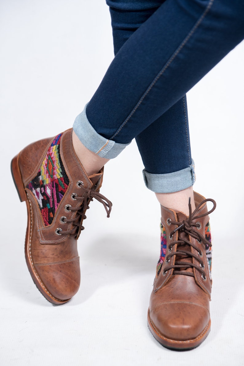 May include: Brown leather ankle boots with colorful woven textile panels. The boots have a lace-up design and are paired with dark blue denim jeans with rolled cuffs. The boots appear to be new.
