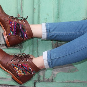 May include: Brown leather ankle boots with colorful woven textile panels. The boots have a square toe, lace-up closure, and a low heel. The boots are worn with blue jeans.