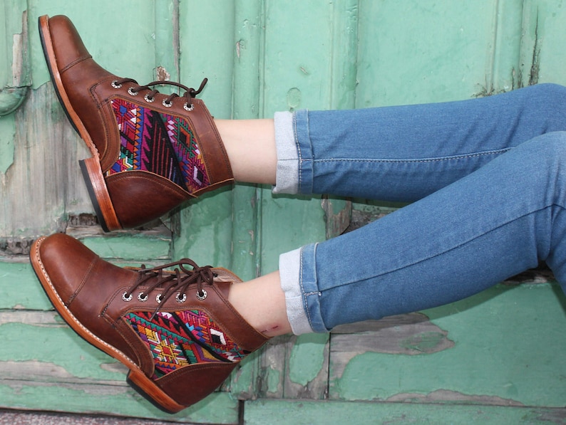May include: Brown leather ankle boots with colorful woven textile panels. The boots have a square toe, lace-up closure, and a low heel. The boots are worn with blue jeans.