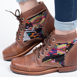 May include: Brown leather ankle boots with colorful woven textile panels. The boots have a lace-up design with metal eyelets and a rounded toe. The textile panels feature geometric patterns in shades of red, yellow, green, and blue. The boots are worn with blue jeans.