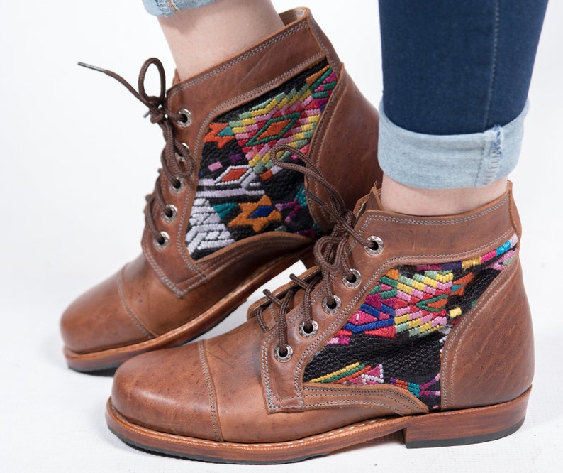 May include: Brown leather ankle boots with colorful woven textile panels. The boots have a lace-up design with metal eyelets and a rounded toe. The textile panels feature geometric patterns in shades of red, yellow, green, and blue. The boots are worn with blue jeans.