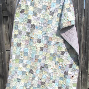 May include: A patchwork quilt featuring a mix of patterned squares in blue, green, grey, and white. The quilt is draped over a wooden fence, highlighting its detailed design and soft texture. The quilt measures approximately 178 cm by 203 cm.