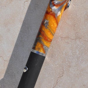 May include: A laser pointer with a matte black lower body and a silver-toned metal button. The upper portion features a swirling, marbled pattern of vibrant orange, metallic gold, and silver. A silver clip is attached to the top cap.
