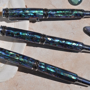 May include: Three luxury fountain pens with iridescent abalone shell barrels and silver-tone accents. The pens are arranged on a light-colored surface, with a seashell and a decorative glass bead nearby. The pens have a sophisticated, elegant design.