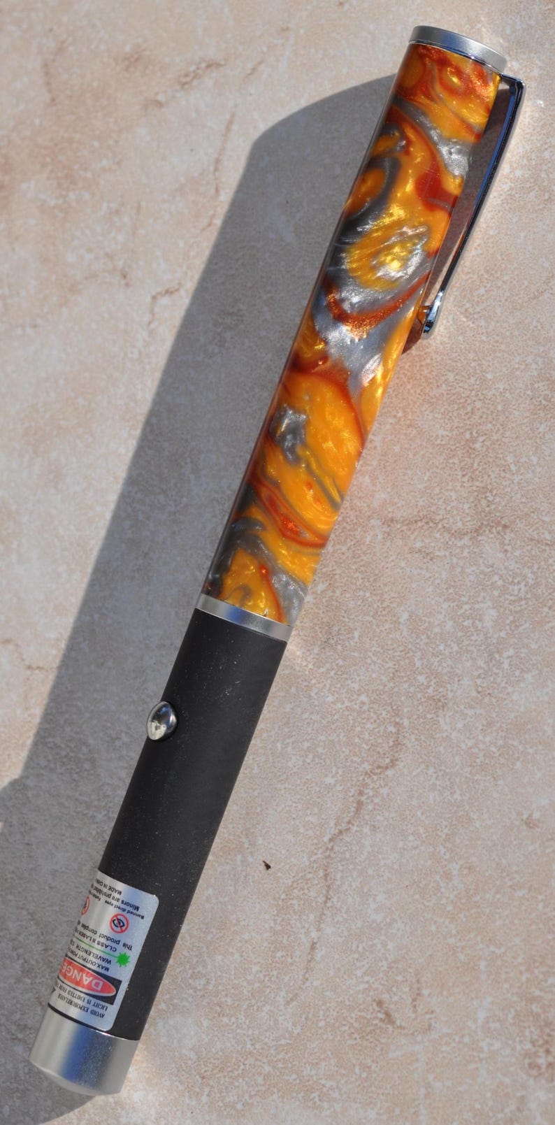 May include: A laser pointer with a matte black lower body and a silver-toned metal button. The upper portion features a swirling, marbled pattern of vibrant orange, metallic gold, and silver. A silver clip is attached to the top cap.