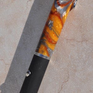 May include: A laser pointer with a matte black lower body and a swirling orange, silver, and gold resin upper barrel. The device features a silver metal pocket clip, a circular silver activation button, and a warning label near the base.