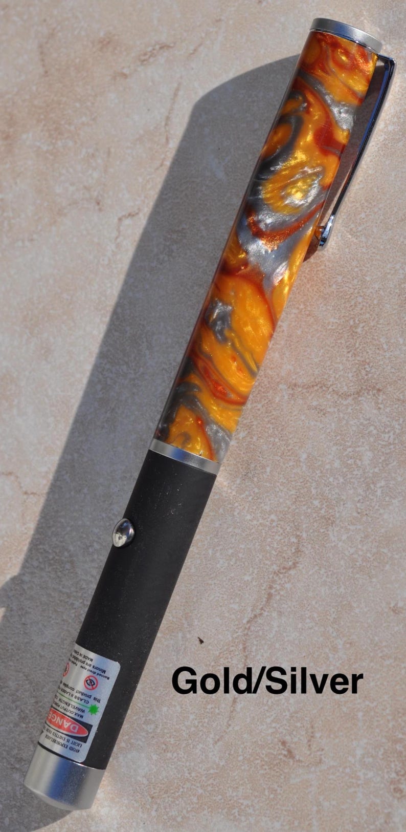 May include: A laser pointer with a matte black lower body and a swirling orange, silver, and gold resin upper barrel. The device features a silver metal pocket clip, a circular silver activation button, and a warning label near the base.