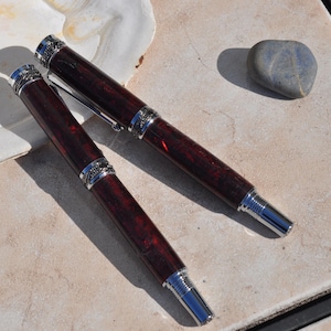 May include: Two stylish pens featuring dark red wooden barrels and silver-coloured detailing. The pens have a classic design with ornate detailing on the caps and mid-sections. The pens are displayed on a light-coloured surface.