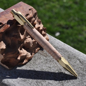 Puede incluir: Un bolígrafo de madera con una punta y un capuchón de metal dorado. El bolígrafo está sobre una pieza de madera con un patrón moteado marrón y gris.