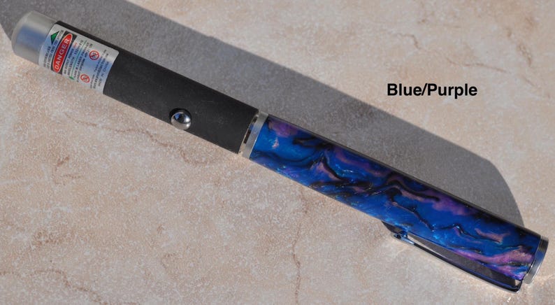May include: A laser pointer with a black matte body, a silver button, and a blue and purple marbled acrylic barrel. The device features a silver tip with a warning label that reads DANGER and includes the text Blue/Purple on the side.