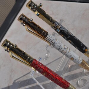 May include: Three bolt-action pens with gold-toned metal hardware and clips. Each pen features a barrel decorated with a printed circuit board pattern in red, white, or black. The pens are displayed on a clear acrylic stand, showing the metallic caps.