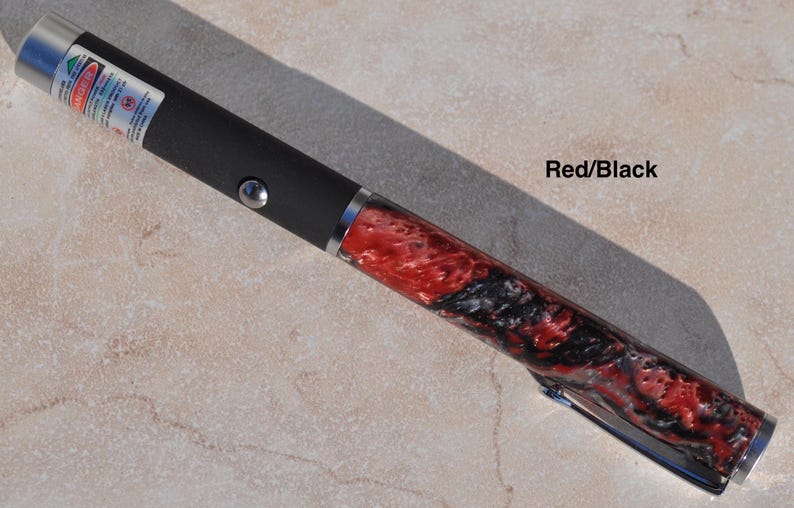May include: A laser pointer featuring a matte black cylindrical body with a silver button and a warning label near the tip. The lower section displays a marbled red and black resin pattern with a silver pocket clip and a polished silver end cap.