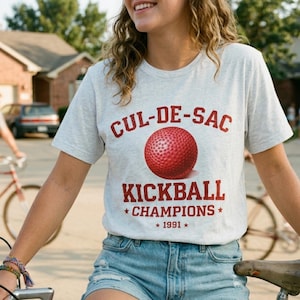 May include: Light gray t-shirt with the text "CUL-DE-SAC KICKBALL CHAMPIONS * 1991 *" in red. A red kickball is centered above the text. The shirt is paired with denim shorts.