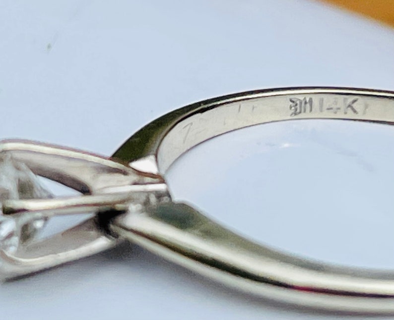 Puede incluir: Un anillo de plata con un solo diamante engastado en garras. La banda est&aacute; inscrita con el texto "14K".