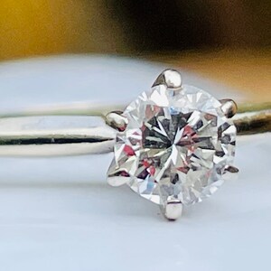 Puede incluir: Un anillo de compromiso de plata con un diamante redondo y transparente.