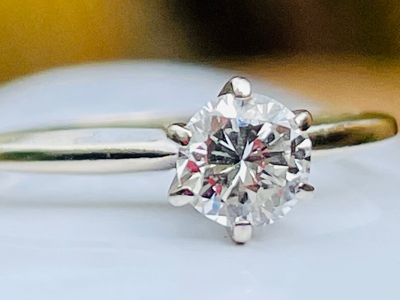 Puede incluir: Un anillo de compromiso de plata con un diamante redondo y transparente.