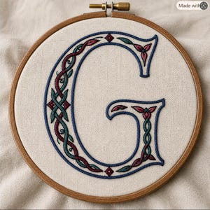 May include: A circular wooden embroidery hoop containing a piece of off-white fabric. The fabric features a large, embroidered capital letter G in dark blue thread, decorated with an intricate, interwoven pattern of teal and maroon thread.