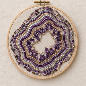 May include: A circular wooden embroidery hoop containing a beaded geode design. The artwork features concentric wavy rows of purple, silver, and gold seed beads surrounding a central cluster of faceted purple and clear crystal-like beads.
