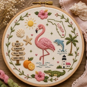 May include: A circular embroidery hoop featuring a Florida-themed design on white fabric. Elements include a pink flamingo, a map of Florida, a dolphin, a lighthouse, an alligator, a sun, a seashell, a starfish, oranges, and a signpost with text.