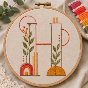 May include: A circular wooden embroidery hoop containing a cream-colored fabric panel with a hand-stitched letter H. The design features green embroidered leaves, orange and pink geometric lines, a small rainbow arc, and circular accents.
