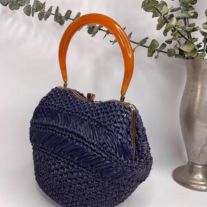 May include: A vintage navy blue woven handbag with an orange handle. The bag has a rounded shape with a gold-colored clasp. The handle is a curved, semi-circular shape. The bag is made of a woven material.