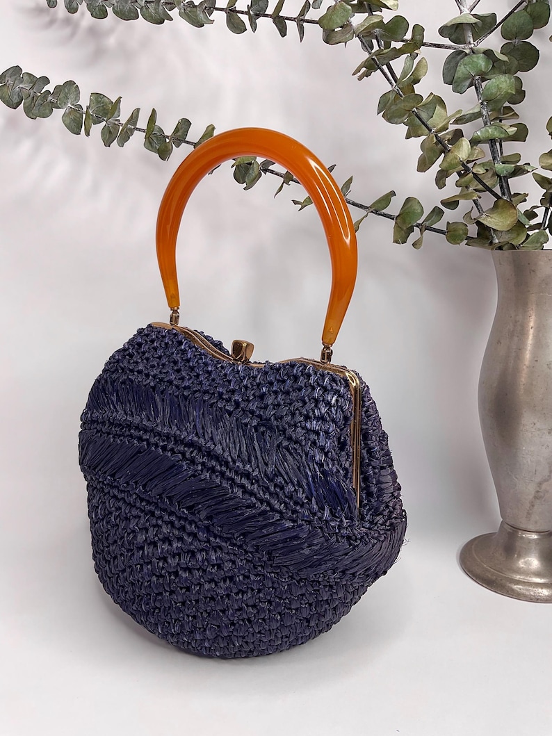 May include: A vintage navy blue woven handbag with an orange handle. The bag has a rounded shape with a gold-colored clasp. The handle is a curved, semi-circular shape. The bag is made of a woven material.