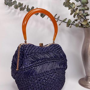 May include: A vintage navy blue woven handbag with a curved amber handle. The bag has a gold-colored clasp and trim. The woven texture creates a unique pattern. The bag is a stylish accessory.