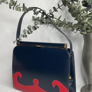 May include: A vintage navy blue handbag with a red wave design on the front. The bag has a rectangular shape, a top handle, and gold-colored hardware. The bag is displayed with a vase of greenery.